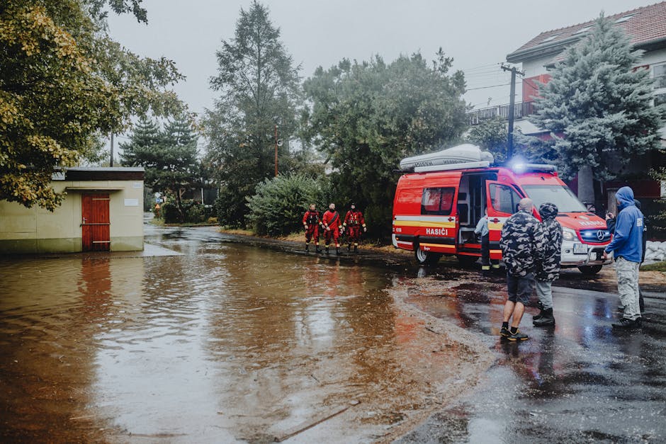A black and white photograph of a residential street affected by flooding, showing a large vehicle parked on the wet, muddy road with water reflecting its image. In the background, several workers in 