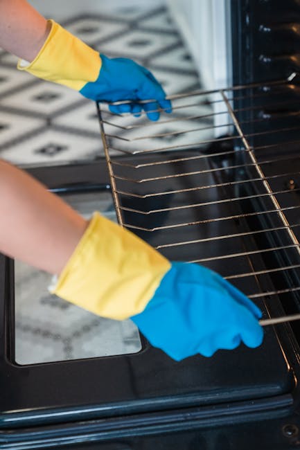 Close-up of a person wearing yellow and blue rubber gloves, using a wire rack inside a modern black oven with a stainless steel interior. The oven door is partially open, revealing the interior cavity