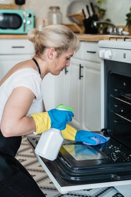 Close-up of a person wearing yellow and blue rubber gloves, using a wire rack inside a modern black oven with a stainless steel interior. The oven door is partially open, revealing the interior cavity