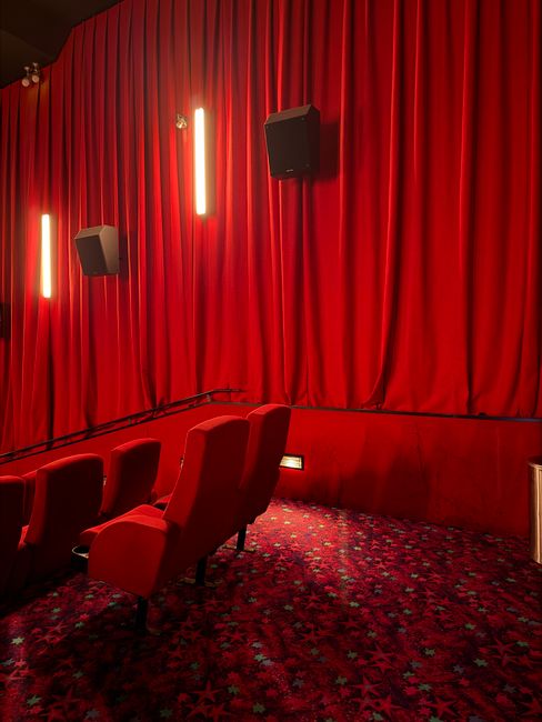 A professional cleaner in protective clothing using a cloth to wipe and sanitise the fabric-covered seats of the Phoenix Cinema, located in Finchley. The seats are arranged in rows within the dimly li