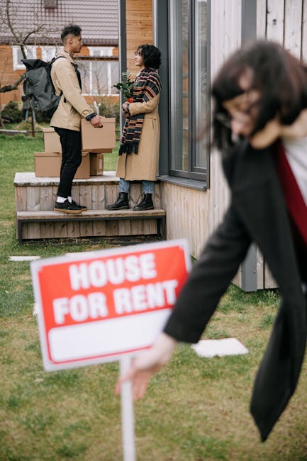 Two individuals, a man and a woman, stand on a wooden porch outside a modern residential building with large glass sliding doors and gray window frames. The man, carrying a backpack, holds a cardboard box, indicating a move or delivery. The woman, dressed in a long coat, has purple hair and appears to engage in conversation with him. In the foreground, a woman dressed in a dark blazer with a red shirt underneath is slightly blurred as she holds a bright red and white 'House For Rent' sign, emphasizing the housing context. The scene is set in a grassy area with a garden in the background, suggesting an outdoor residential environment during daylight hours. The interior of the house is not visible, but the exterior elements and the movement imply a transition or rental process, fitting the context of housing and property management.